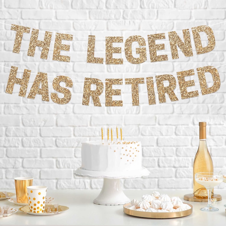 Gold glitter banner with the text "The Legend Has Retired" hanging on a white brick wall. A white cake with gold sprinkles sits on a white cake stand in front of the banner. There are gold and white party supplies on the table.