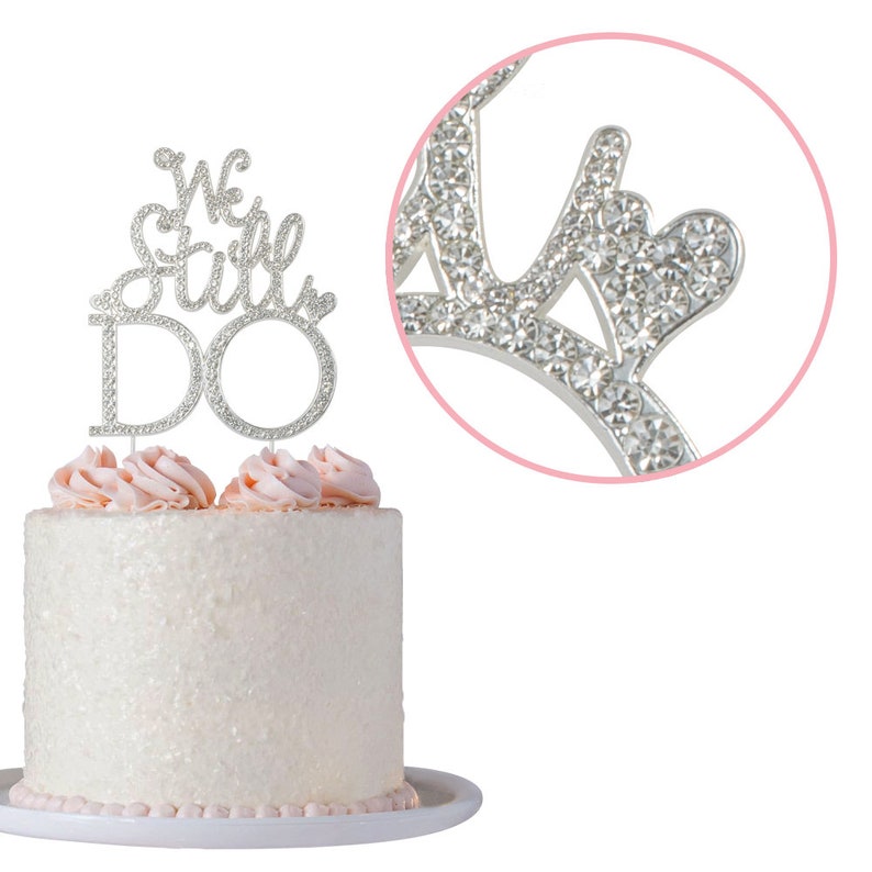 May include: A white cake with pink frosting and a silver cake topper that reads "We Still Do." The cake topper is encrusted with rhinestones. The cake sits on a white plate.