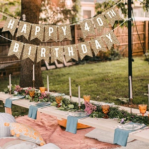 A gold glitter banner with the text "Happy 40th Birthday" hanging in a backyard setting. A long wooden table is set with a floral centerpiece, blue napkins, and amber glasses. The table is surrounded by pillows and blankets.