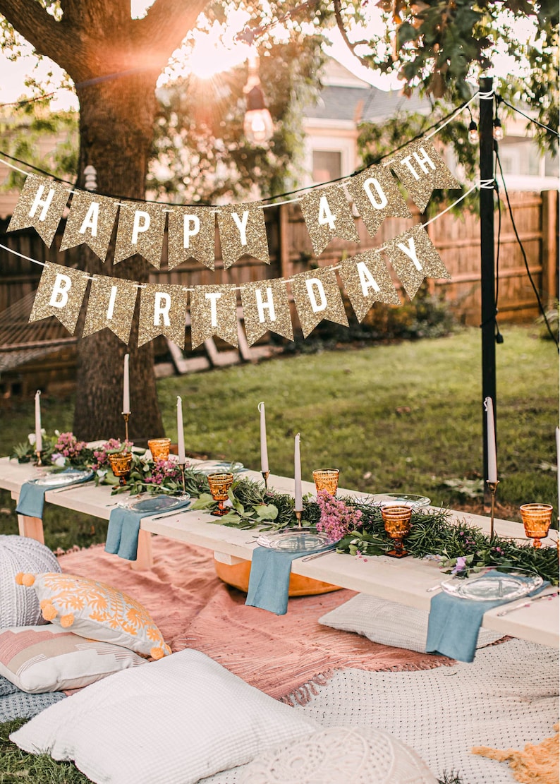A gold glitter banner with the text "Happy 40th Birthday" hanging in a backyard setting. A long wooden table is set with a floral centerpiece, blue napkins, and amber glasses. The table is surrounded by pillows and blankets.