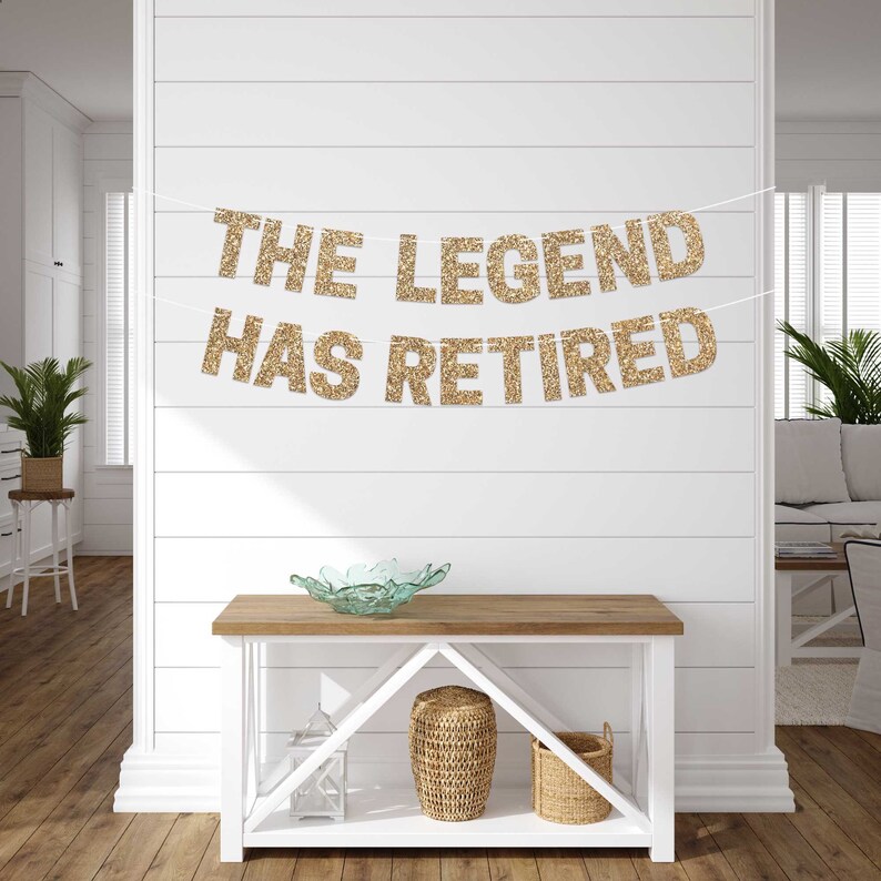 Gold glitter banner with the text "The Legend Has Retired". The banner is hanging on a white wall with wood paneling. A wooden table with a glass bowl and two wicker baskets is in the foreground.