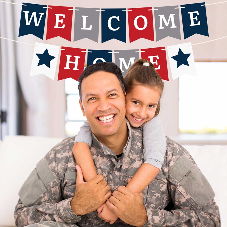 A father in military uniform smiles and hugs his daughter. They are standing in front of a white couch with a red, white, and blue banner that says "Welcome Home".