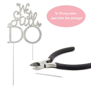 May include: A silver cake topper with the words "We Still Do" encrusted with rhinestones. The topper has two prongs for insertion into a cake. A pair of black-handled pliers and a cut prong are also in the image.