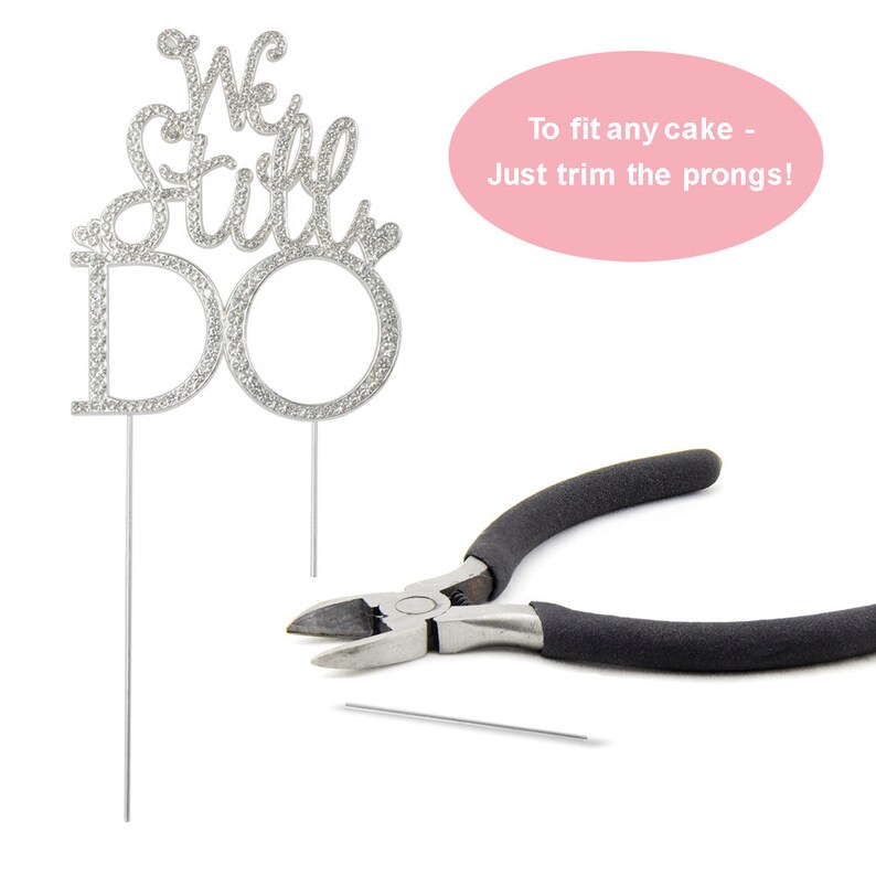 May include: A silver cake topper with the words "We Still Do" encrusted with rhinestones. The topper has two prongs for insertion into a cake. A pair of black-handled pliers and a cut prong are also in the image.