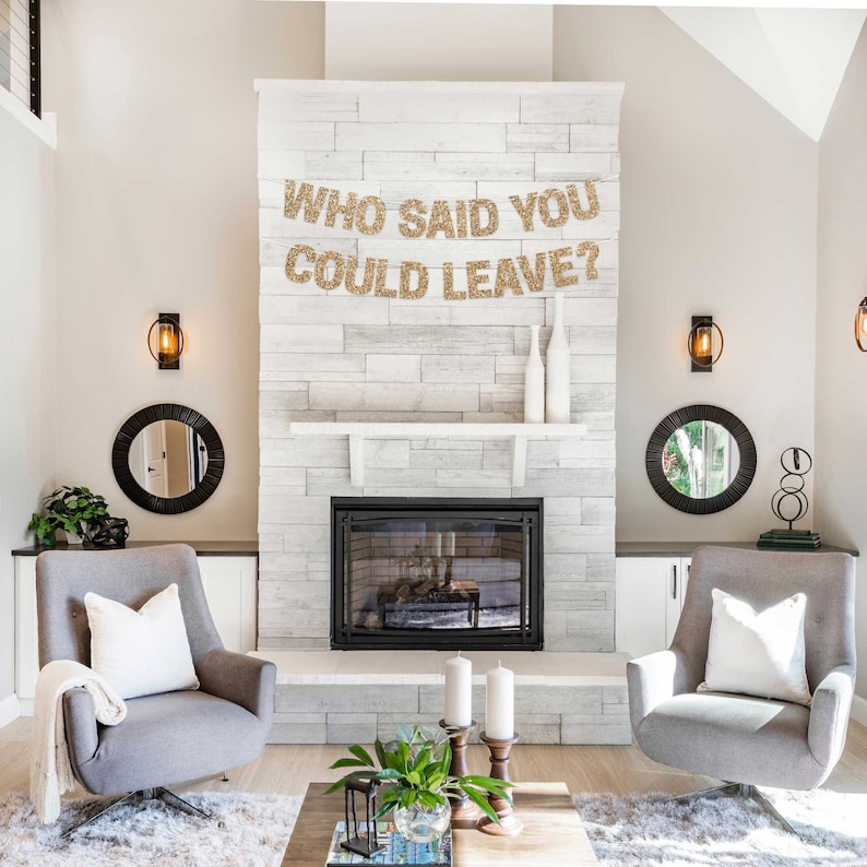 Gold glitter banner with the text "Who said you could leave?" hanging above a fireplace in a living room.