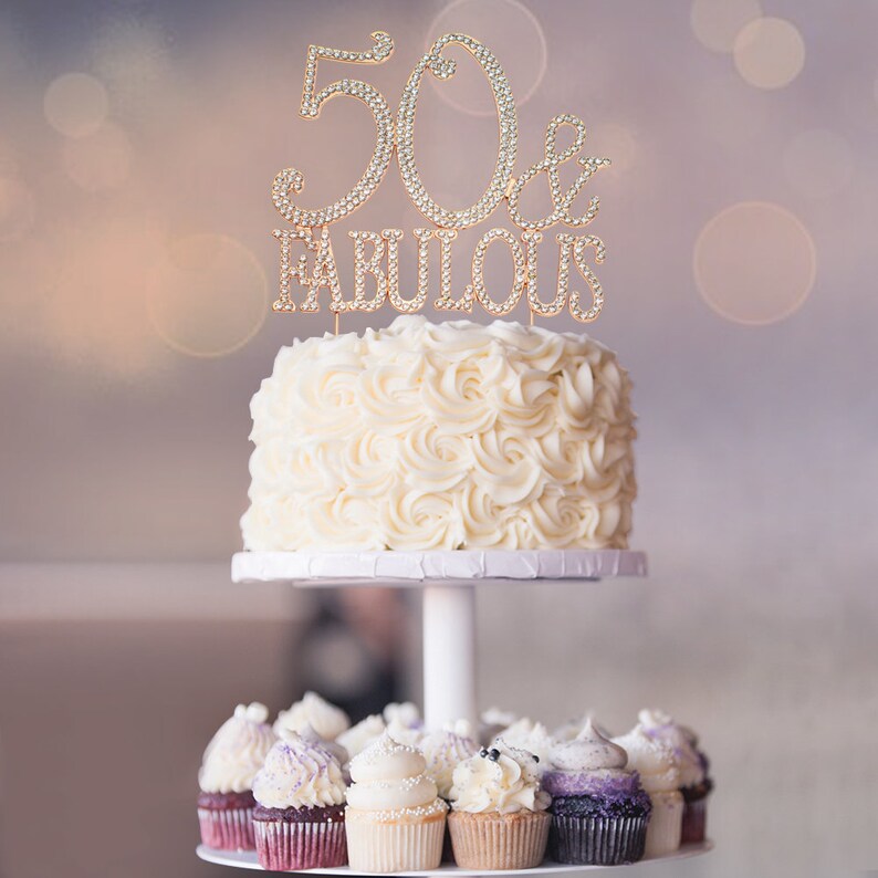 May include: A white cake with white frosting and a gold '50 & Fabulous' cake topper. The cake is on a white cake stand with cupcakes on the bottom tier.