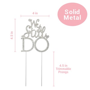 May include: A silver metal cake topper that reads "We Still Do" with rhinestone accents. The topper is 4 inches wide and 4.5 inches tall, with 4.5-inch trimmable prongs. The words are in a script font. The topper is for a wedding or anniversary.
