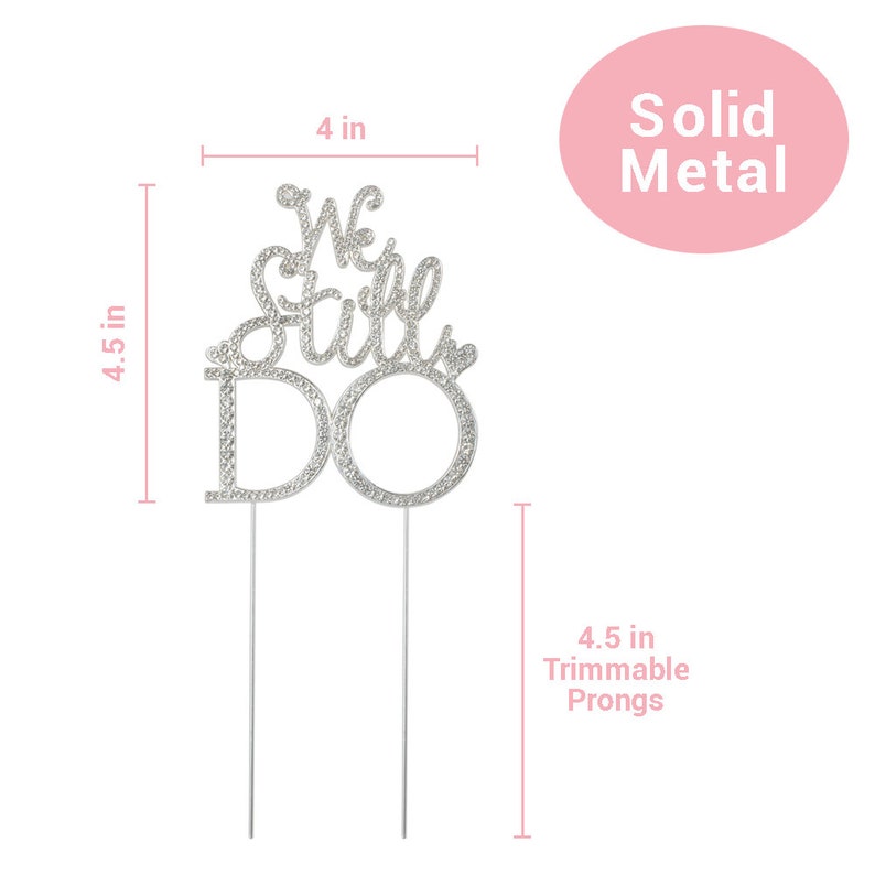 May include: A silver metal cake topper that reads "We Still Do" with rhinestone accents. The topper is 4 inches wide and 4.5 inches tall, with 4.5-inch trimmable prongs. The words are in a script font. The topper is for a wedding or anniversary.