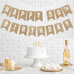 Gold glitter banner with the text "Happy 50th Birthday". The banner is hanging on a white brick wall. There is a white cake with gold sprinkles on a white cake stand, a bottle of wine, and a plate of white meringue cookies on a gold tray.