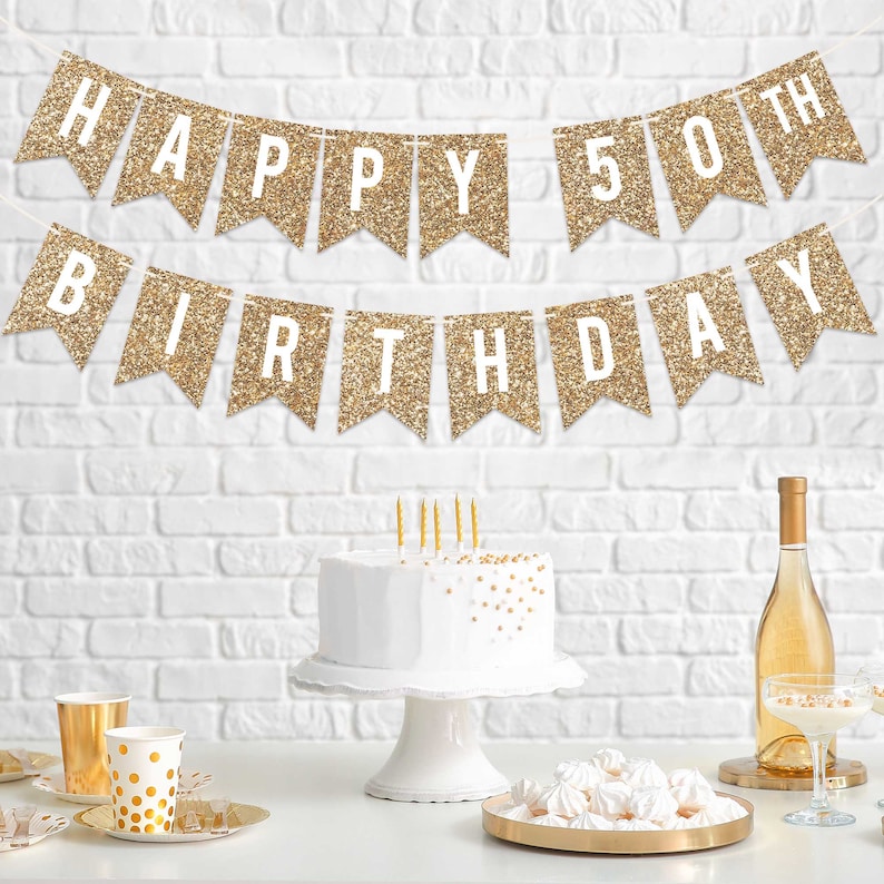 Gold glitter banner with the text "Happy 50th Birthday". The banner is hanging on a white brick wall. There is a white cake with gold sprinkles on a white cake stand, a bottle of wine, and a plate of white meringue cookies on a gold tray.