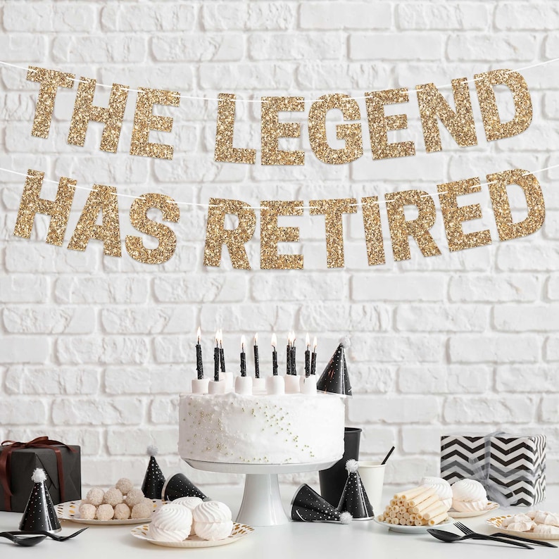 Gold glitter banner with the text "The Legend Has Retired" hanging on a white brick wall. A white cake with black candles is on a white cake stand in front of the banner. There are black party hats, white and black striped paper straws, and white and black patterned gift boxes on a white table in front of the cake.