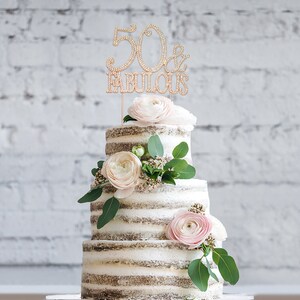 May include: A three-tiered cake decorated with white and pink flowers and greenery. The top tier has a gold cake topper that says "50 & Fabulous".