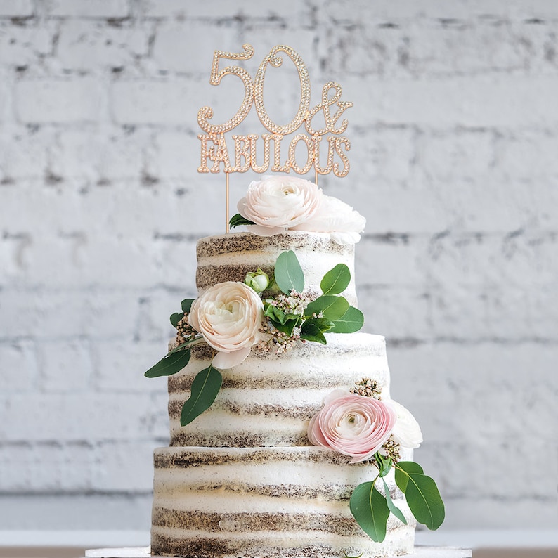 May include: A three-tiered cake decorated with white and pink flowers and greenery. The top tier has a gold cake topper that says "50 & Fabulous".