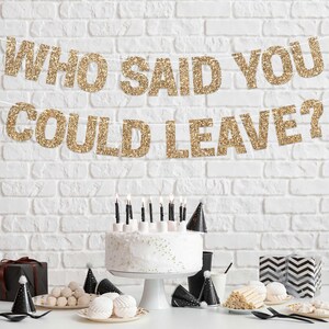 Gold glitter banner with the text "Who said you could leave?" hanging above a white cake with black candles and black party hats on a table with black and white decorations.