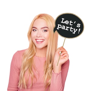 May include: A woman holds a black and gold glitter speech bubble photo booth prop that says "Let's party!"