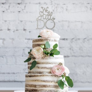 May include: A three-tiered wedding cake with a "We Still Do" cake topper. The cake is decorated with light pink flowers and green foliage. The cake has a rustic, semi-naked design.