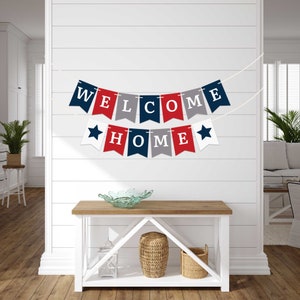 A red, white, and blue banner with the words "WELCOME HOME" hanging on a white wall. The banner is made of felt and has stars on each end.