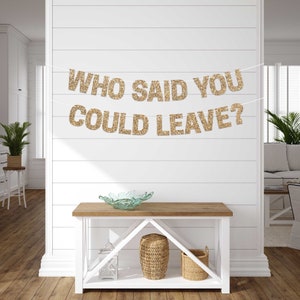 Gold glitter banner with the text "Who said you could leave?" hanging on a white wall. The banner is strung with white thread and is hanging above a wooden table with a glass bowl on top.