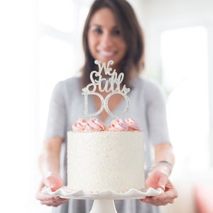 May include: A white frosted cake with pink frosting and a silver cake topper that reads "We Still DO". The cake is on a white pedestal plate.