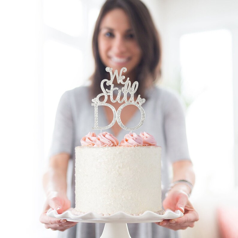 May include: A white frosted cake with pink frosting and a silver cake topper that reads "We Still DO". The cake is on a white pedestal plate.