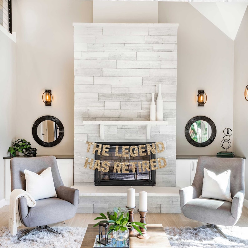 A gold glitter banner with the text "The Legend Has Retired" hanging above a fireplace in a living room. The banner is strung with white twine and has gold glitter letters.