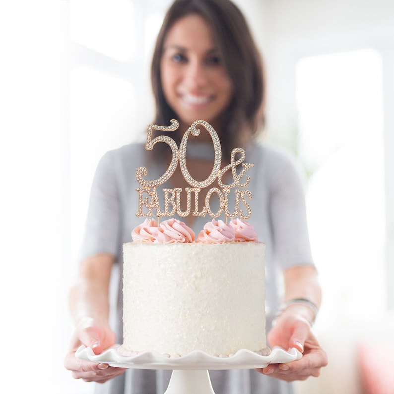 May include: A gold cake topper with rhinestones that says "50 & Fabulous" on a white cake with pink frosting.