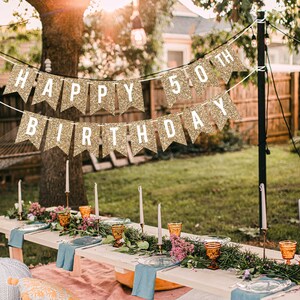 Gold glitter banner with the text "Happy 50th Birthday" hanging in a backyard setting. The banner is strung between two trees and is decorated with white letters.