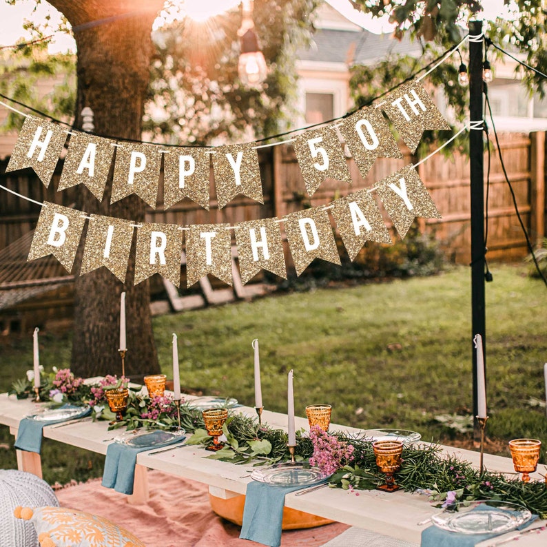 Gold glitter banner with the text "Happy 50th Birthday" hanging in a backyard setting. The banner is strung between two trees and is decorated with white letters.