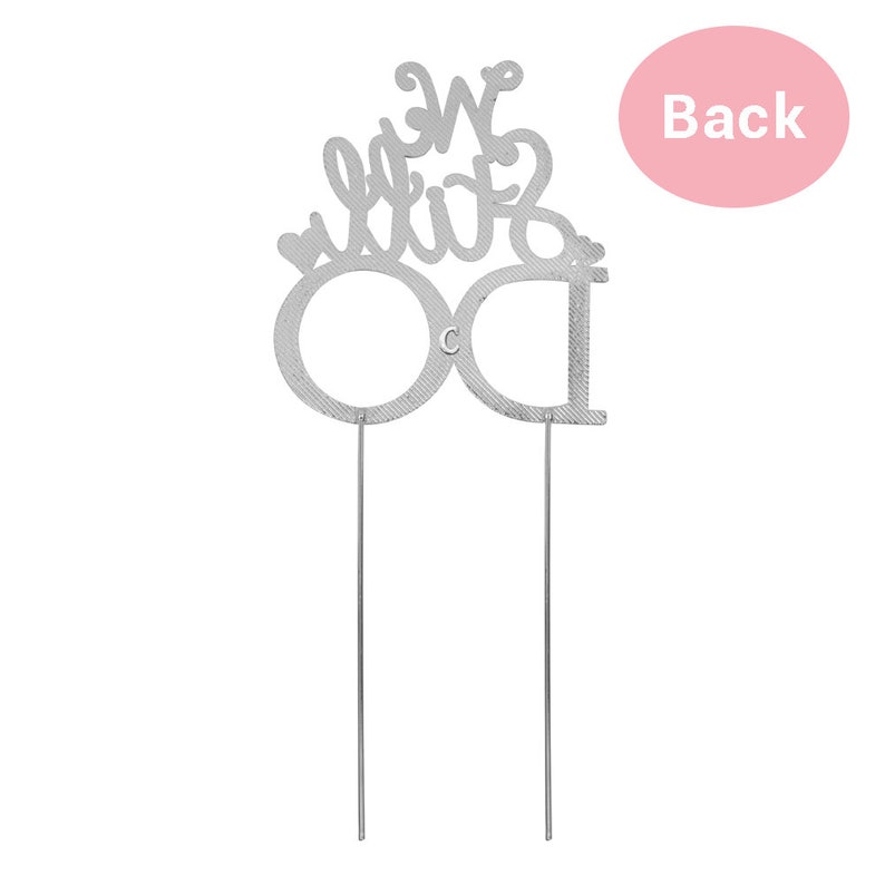 May include: A silver-colored cake topper with the words "We Do" in a decorative script. The letters "O" and "D" are large and stylized. Two thin metal rods extend from the bottom for insertion into a cake. A pink circle with the word "Back" is in the background.