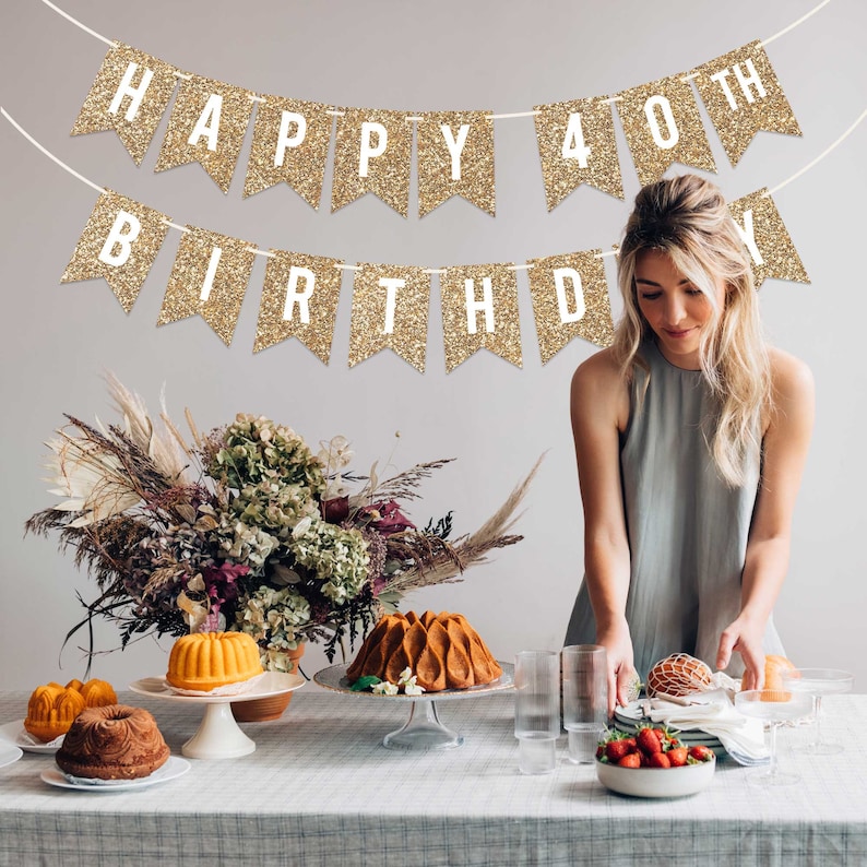 Gold glitter banner with the text "Happy 40th Birthday" hanging above a table with cakes and flowers.