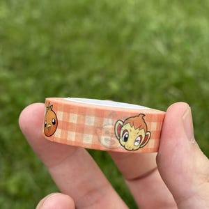 Op de afbeelding: Washi-tape met een oranje en wit geruit patroon, versierd met stripfiguren. De tape wordt in een hand gehouden tegen een groene achtergrond. Decoratief knutselmateriaal.
