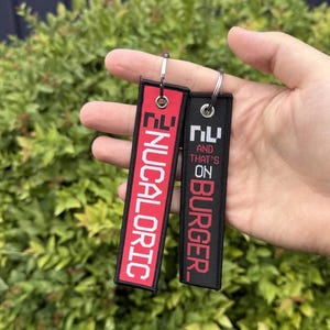 May include: Two rectangular keychains. One is red with the word "NUCALORIC" in white. The other is black with the text "NU AND THAT'S ON BURGER" in red. Both have silver key rings.