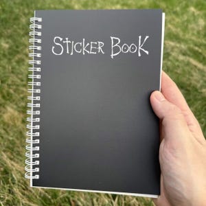 May include: A black spiral-bound sticker book held in a hand. The cover features the words "Sticker Book" in white, handwritten-style lettering. The book's pages are white, and the background is blurred green grass.