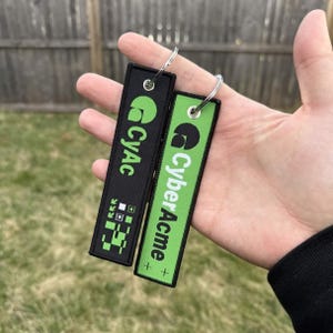 May include: Two rectangular keychains with silver rings. One is black with "CyAc" and a pixelated design in green. The other is green with "CyberAcme" in white. Both have a green logo and are held in a hand.