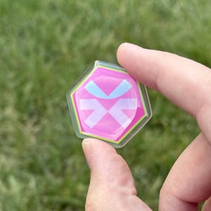 May include: A pink, hexagonal-shaped pin with a white and light blue design. The pin has a clear border with a green outline. The background is blurred green grass.