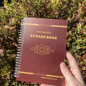 Könnte beinhalten: Ein rotes, spiralgebundenes Stickerbuch mit einem goldenen Vault-Tec-Logo und dem Text "RESTRICTED VAULT DWELLER'S STICKER BOOK RESTRICTED VDSB VTB-001-23"