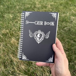 Peut inclure: Un carnet d'autocollants noir à spirale avec du texte blanc et des motifs décoratifs d'angle. La couverture présente les mots "STICKER BOOK" et un motif en forme de cœur avec un crâne et des ailes.