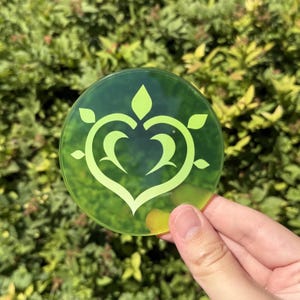 Op de afbeelding: Een ronde, transparante groene onderzetter met een lichtgroen hartvormig ontwerp met bladeren. De onderzetter wordt vastgehouden voor een vage groene, bladerrijke achtergrond. Het ontwerp is een gestileerd bloemenembleem.