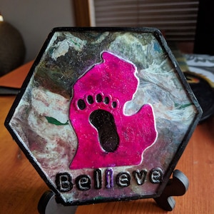 May include: A hexagonal resin art piece with a pink outline of the state of Michigan with a black footprint inside. The piece is on a black stand and has the word "Believe" written on the bottom.