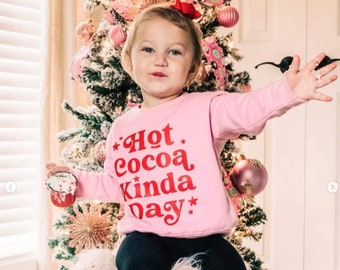 Può includere: Una bambina indossa una felpa rosa con la scritta rossa "Hot Cocoa Kinda Day", abbinata a leggings neri e calzini a righe. La bambina tiene in mano una tazza decorativa davanti a un albero di Natale decorato.