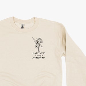Könnte beinhalten: Ein beiger Pullover mit einem Blumendesign und dem Text "Happiness is being a mama" auf der Vorderseite.