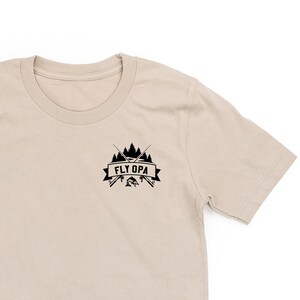 Peut inclure: Un t-shirt à manches courtes beige avec un motif noir de cannes à pêche croisées, un poisson et une bannière qui dit "Fly Opa".
