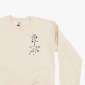 Op de afbeelding: Een beige sweatshirt met een zwarte afbeelding van een bloemenboeket en de tekst "Happiness is being a yoga" op de voorkant.