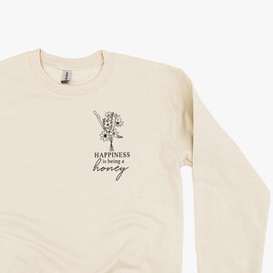 Könnte beinhalten: Ein beiger Pullover mit einem schwarzen Blumendesign und dem Text "Happiness is being a honey" auf der Vorderseite.