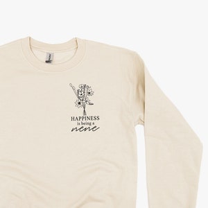 Op de afbeelding: Crèmekleurige sweatshirt met lange mouwen. Een zwart bloemmotief en de tekst "Happiness is being a nene" zijn op de voorkant gedrukt. De sweatshirt is gemaakt van een zacht materiaal.