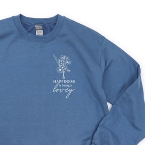 Puede incluir: Una sudadera azul con un gráfico blanco de flores y el texto "Happiness is being a lovey".
