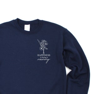 Op de afbeelding: Navyblauwe trui met een wit bloemenmotief en de tekst "Happiness is being a nanny".