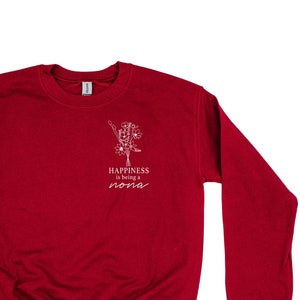 Op de afbeelding: Rood sweatshirt met een wit grafisch ontwerp met de tekst "Happiness is being a Nona" met een bloemillustratie.