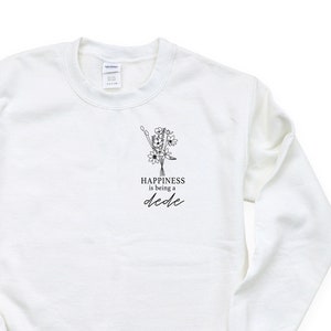Könnte beinhalten: Weißer Pullover mit einer schwarz-weißen Illustration einer Nadel und eines Fadens mit Blumen. Der Text "Happiness is being a dcdlc" ist unter der Illustration gedruckt.