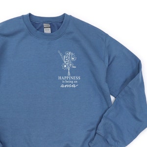 Könnte beinhalten: Blaues Sweatshirt mit dem Text "Happiness is being an ama" und einer weißen Blumengrafik. Das Sweatshirt mit Rundhalsausschnitt ist aus weichem Material.